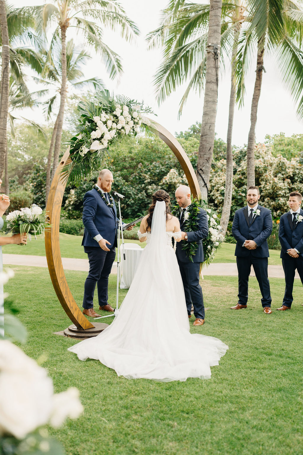 A bride and groom stand facing each other holding hands under a circular floral arch, with an officiant in the background. Palm trees and greenery surround the scene for a wedding at the four seasons in oahu