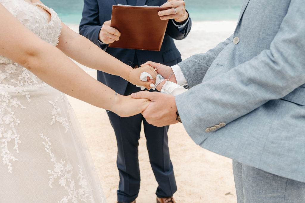 A couple holding hands during a beach wedding ceremony, with an officiant standing behind holding a brown folder for a beach wedding in oahu