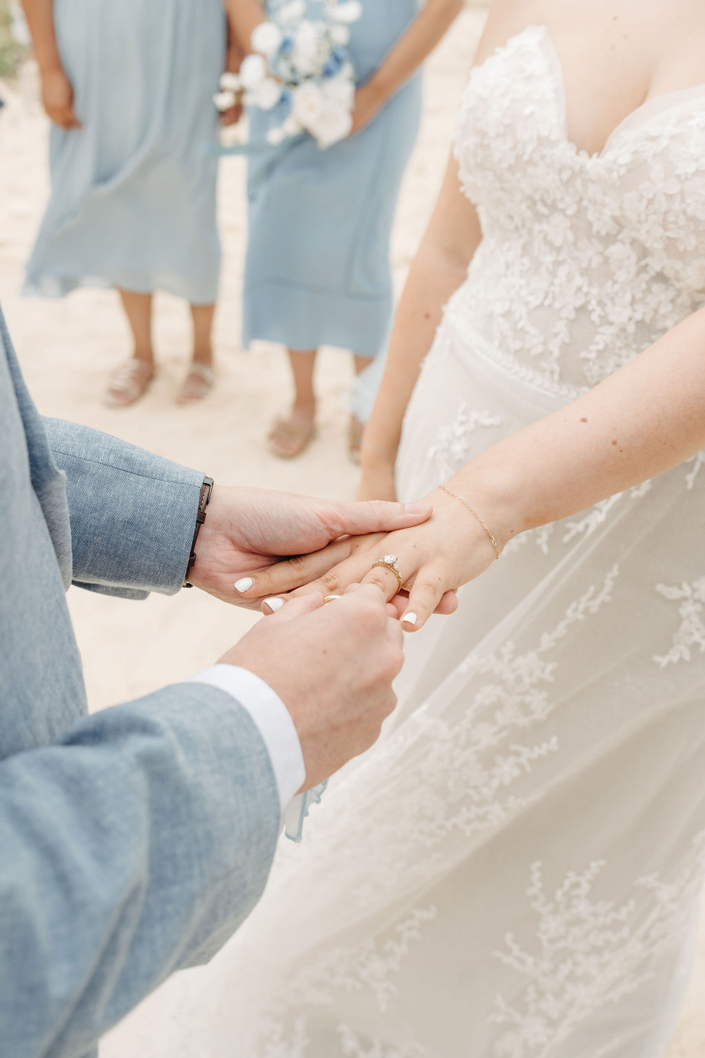 Close-up of a groom placing a ring on the bride's finger during a wedding ceremony. Bridesmaids in blue dresses are visible in the background.