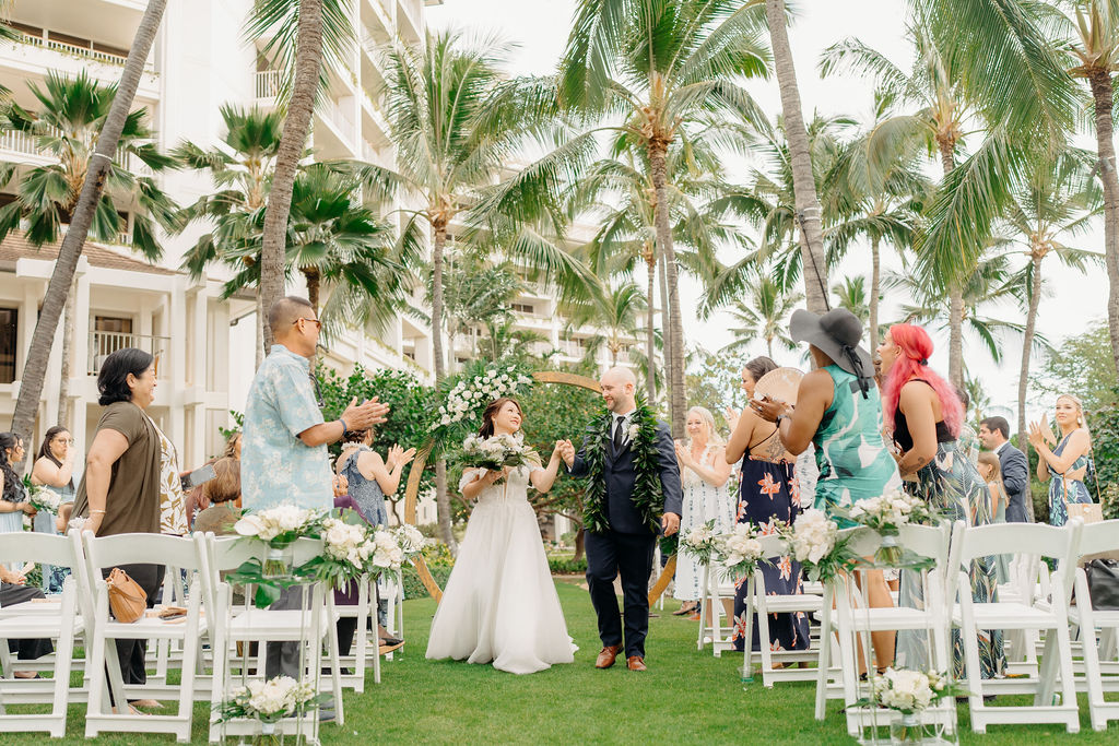 A bride and groom stand facing each other holding hands under a circular floral arch, with an officiant in the background. Palm trees and greenery surround the scene for a wedding at the four seasons in oahu