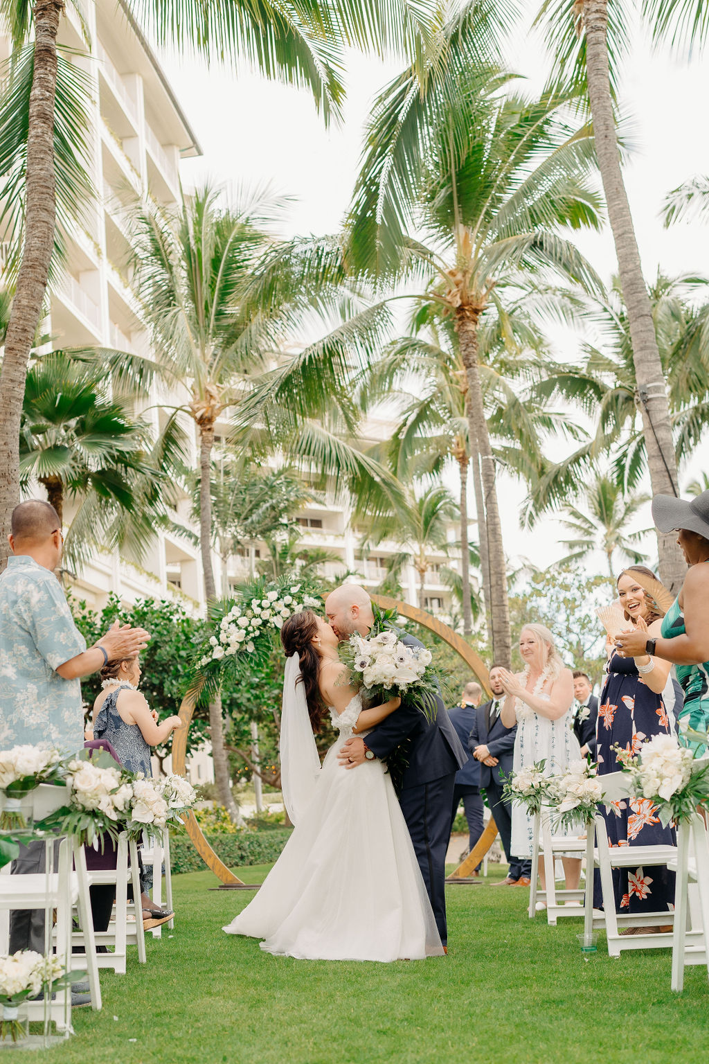 A bride and groom stand facing each other holding hands under a circular floral arch, with an officiant in the background. Palm trees and greenery surround the scene for a wedding at the four seasons in oahu