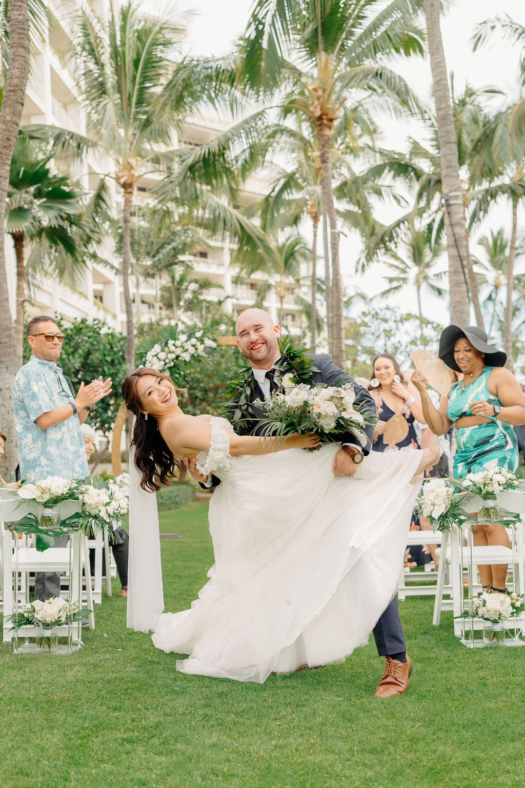 A bride and groom stand facing each other holding hands under a circular floral arch, with an officiant in the background. Palm trees and greenery surround the scene for a wedding at the four seasons in oahu