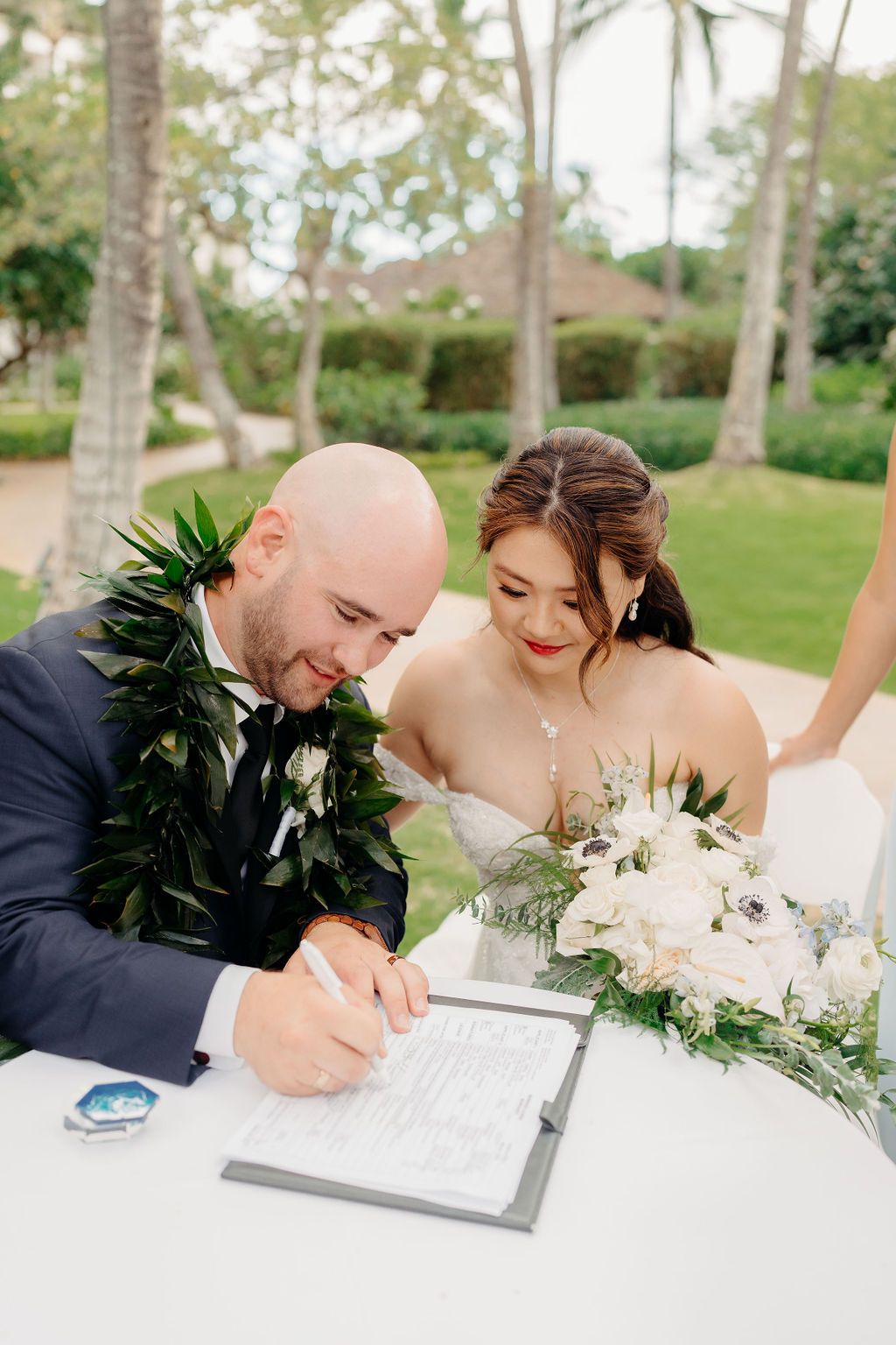 A couple signs a document at an outdoor table surrounded by lush greenery. The man wears a dark suit and lei, while the woman wears a white dress and holds a bouquet.