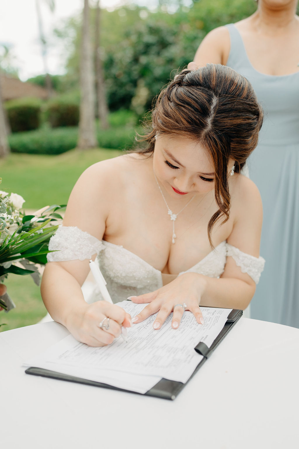 A couple signs a document at an outdoor table surrounded by lush greenery. The man wears a dark suit and lei, while the woman wears a white dress and holds a bouquet.