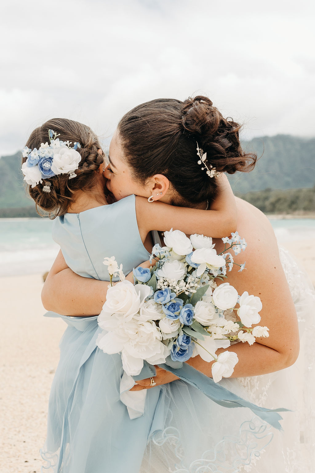 A bride holding a young girl on a beach. The girl wears a blue dress with blue and white flowers in her hair. The bride holds a bouquet of blue and white flowers.