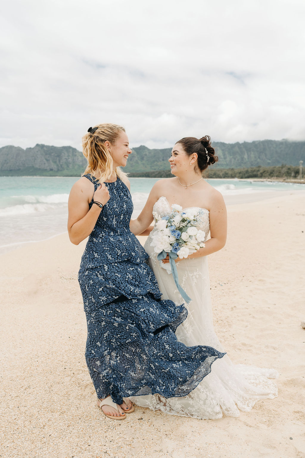 Two women in formal dresses stand on a beach. One wears a blue dress, the other a white gown holding a bouquet. They are smiling at each other with the ocean and mountains in the background.