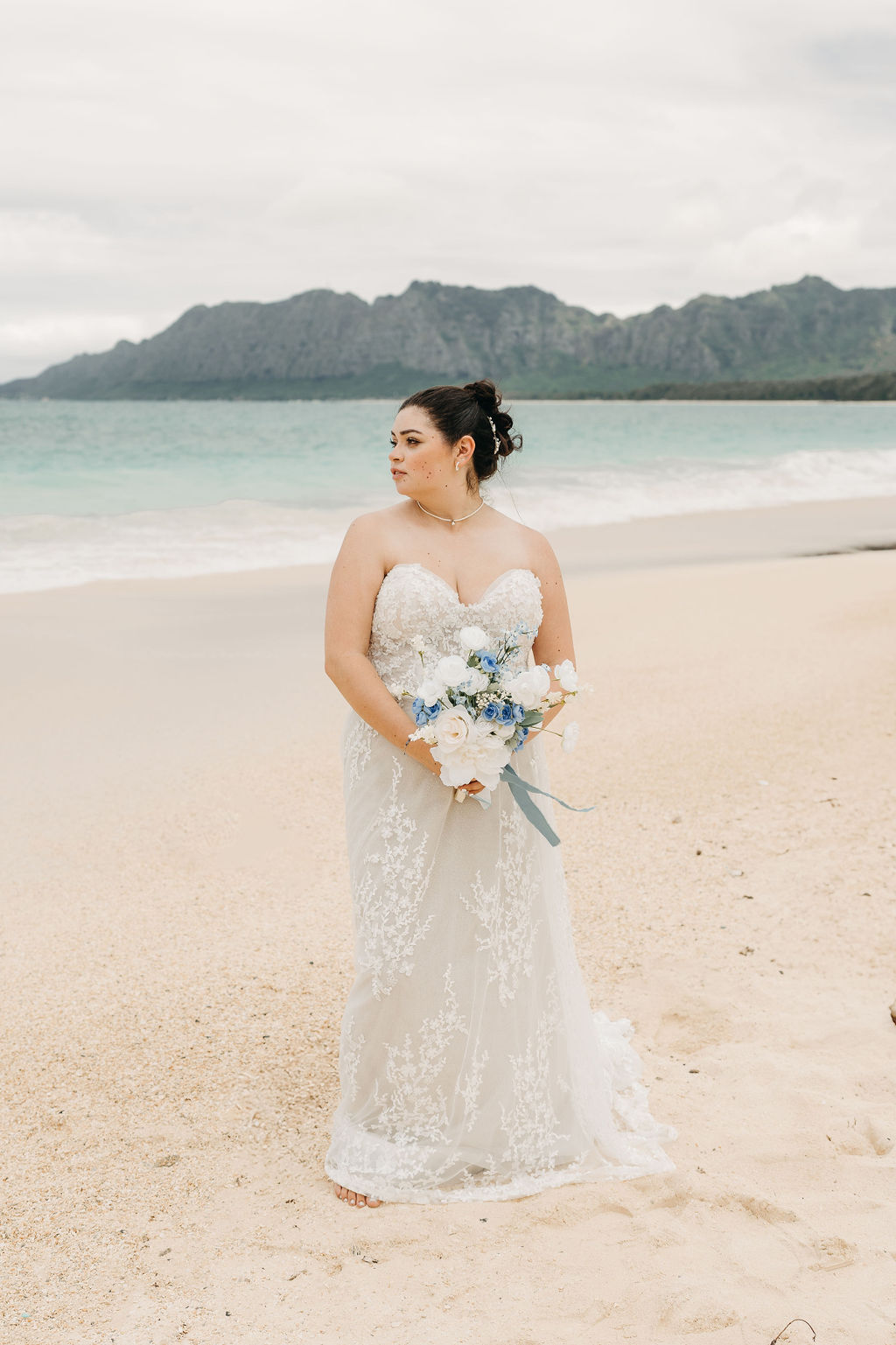 Bride in a strapless lace wedding dress holding a bouquet, standing on a beach with mountains and ocean in the background.