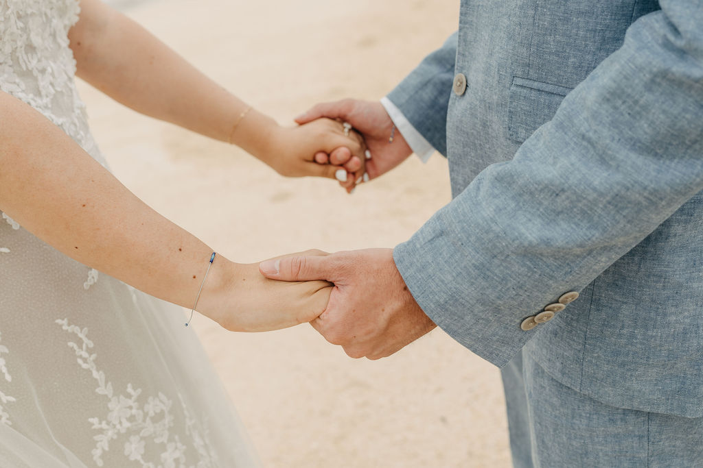 A bride and groom holding hands, showing part of the wedding dress and suit, standing on a sandy surface.