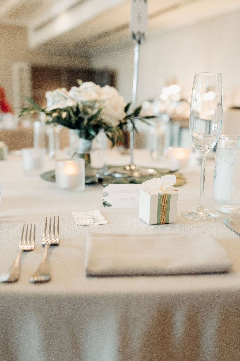 A table set for an event, adorned with white flowers and greenery. Plates, cutlery, and glasses are neatly arranged. In the background, people sit at table sat the four seasons in oahu