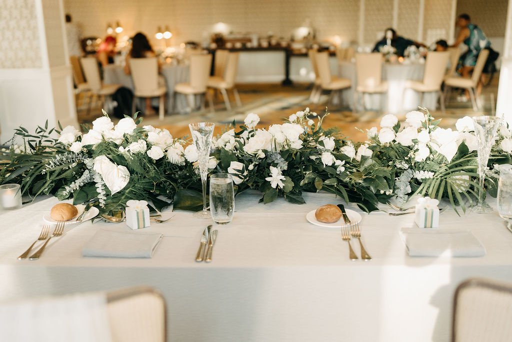 A table set for an event, adorned with white flowers and greenery. Plates, cutlery, and glasses are neatly arranged. In the background, people sit at table sat the four seasons in oahu