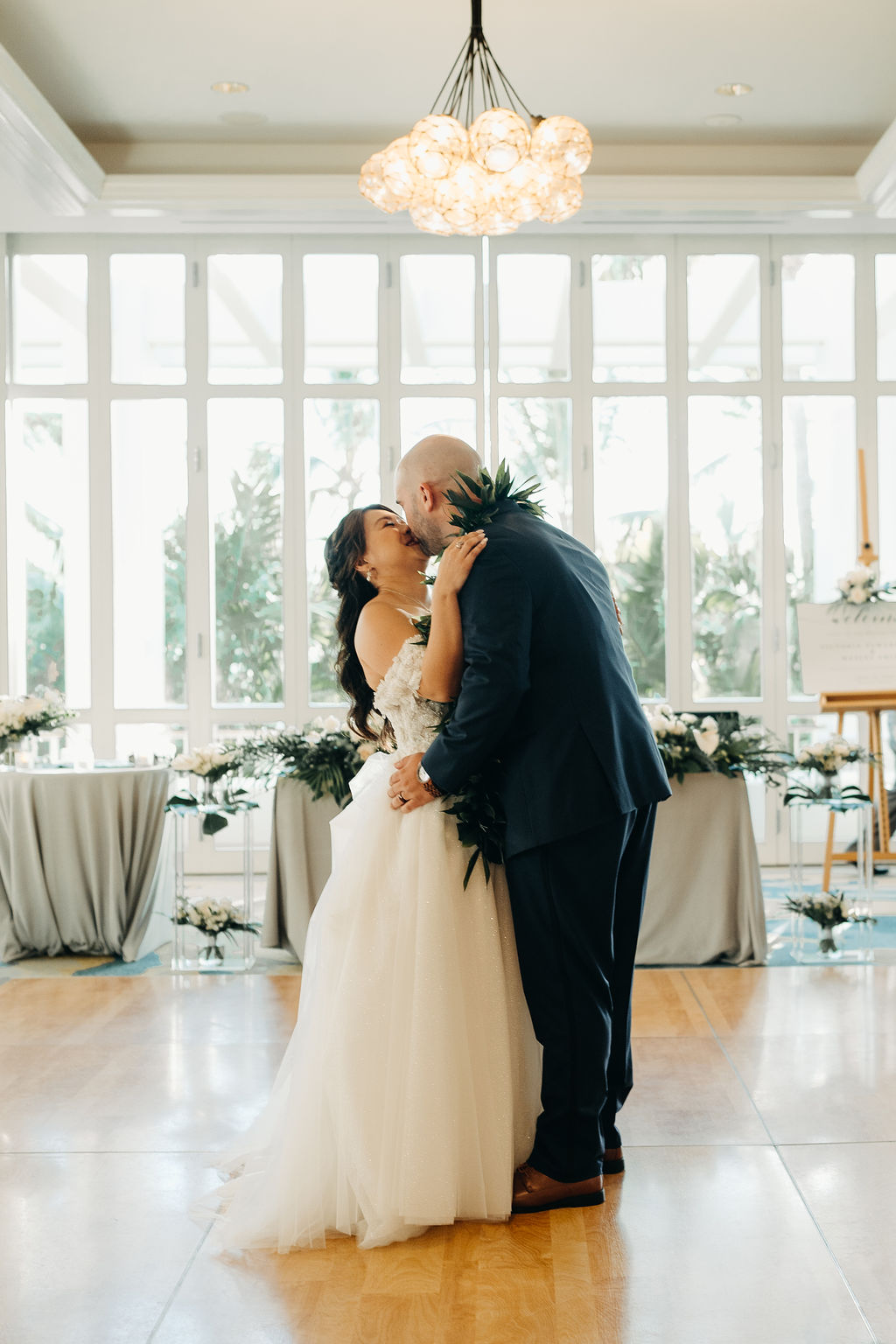 A bride and groom kiss on a dance floor in a well-lit room, with white flowers and draped tables in the background for a wedding at the four seasons in Oahu