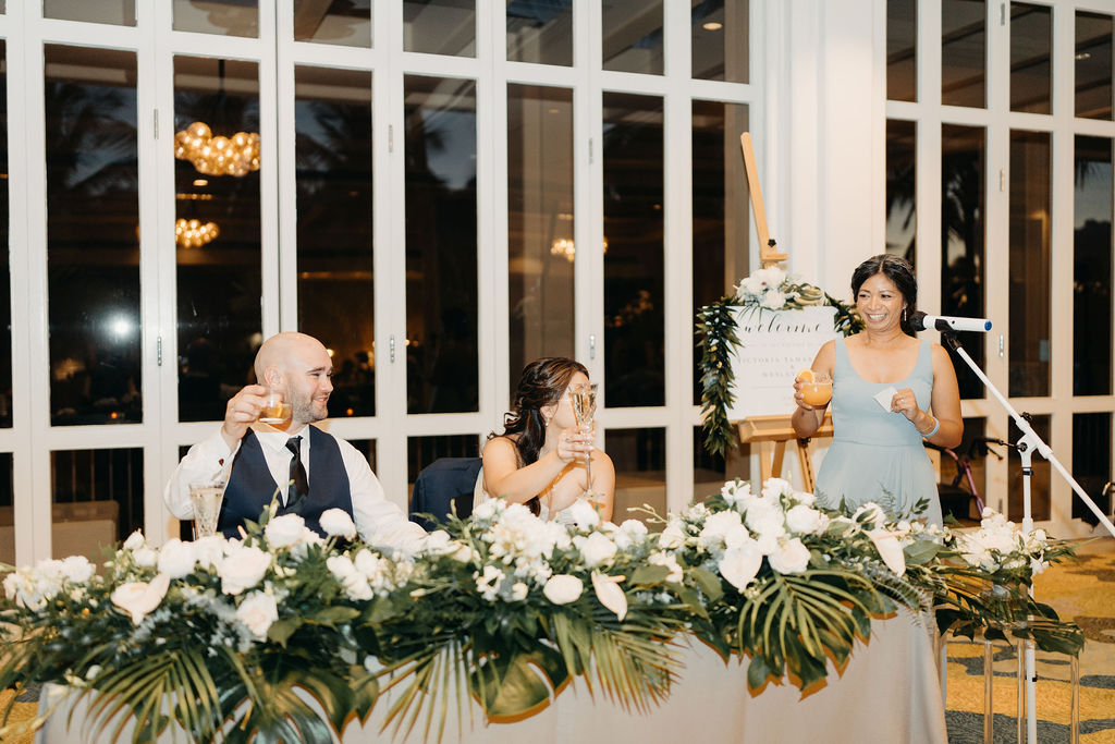 Three people at a decorated table, raising glasses in a toast. A woman stands at a microphone, while the others sit. Floral arrangements adorn the table.