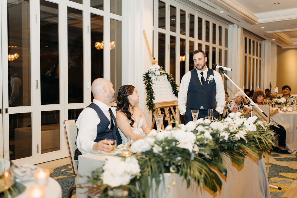 Three people at a decorated table, raising glasses in a toast. A woman stands at a microphone, while the others sit. Floral arrangements adorn the table.