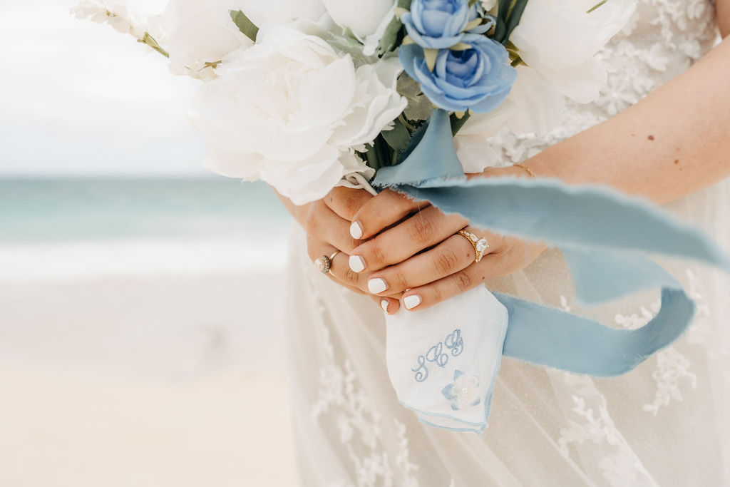 A bride holds a bouquet of white and blue flowers with a blue ribbon, standing on a beach.