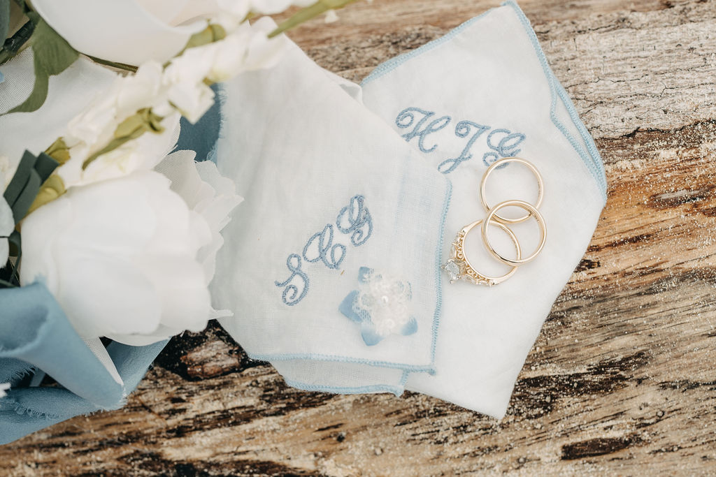 Two monogrammed handkerchiefs with floral embroidery are placed on a wooden surface next to three gold rings.