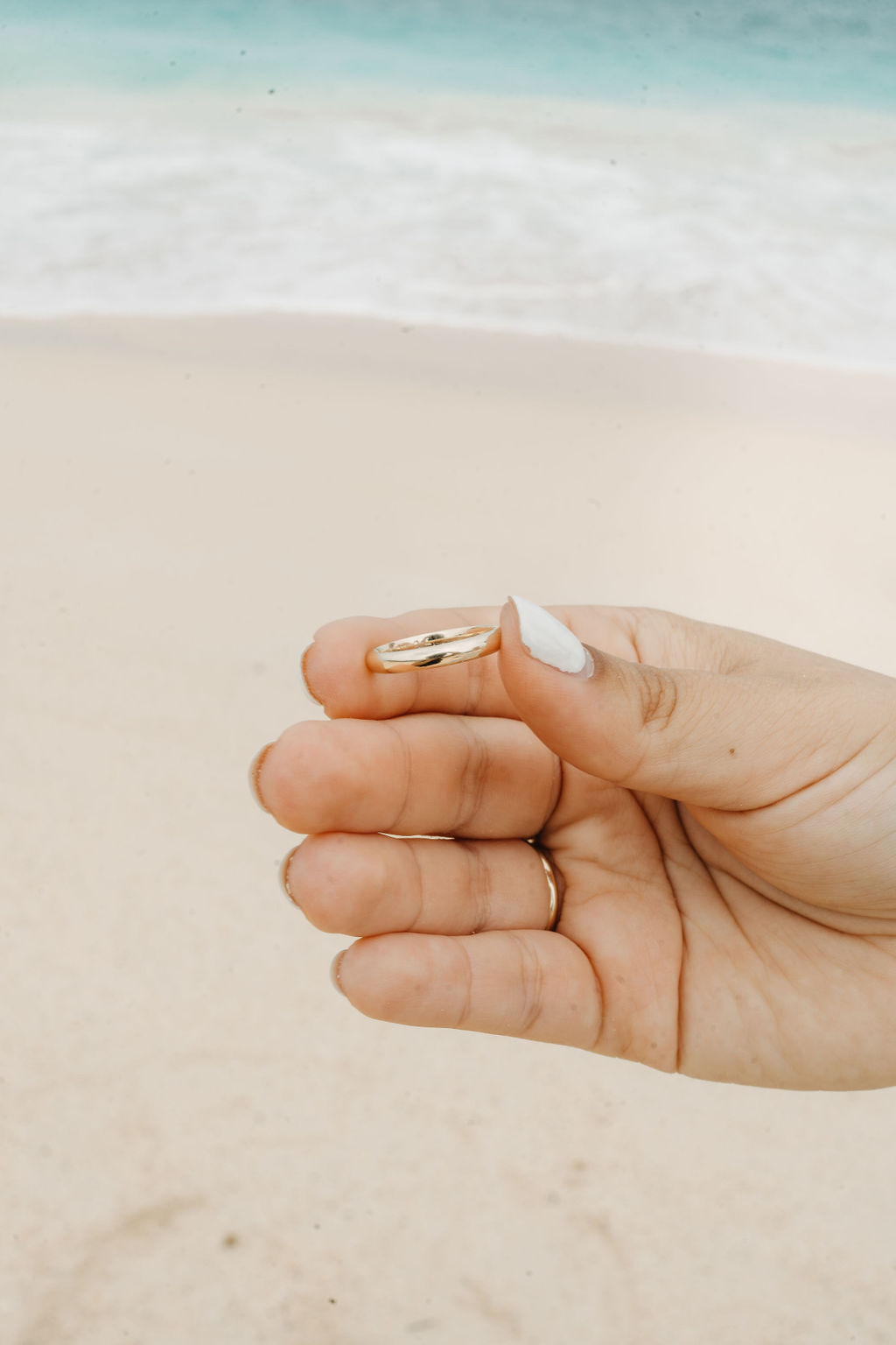 A hand with white nail polish holds a gold ring against a sandy beach background with waves.