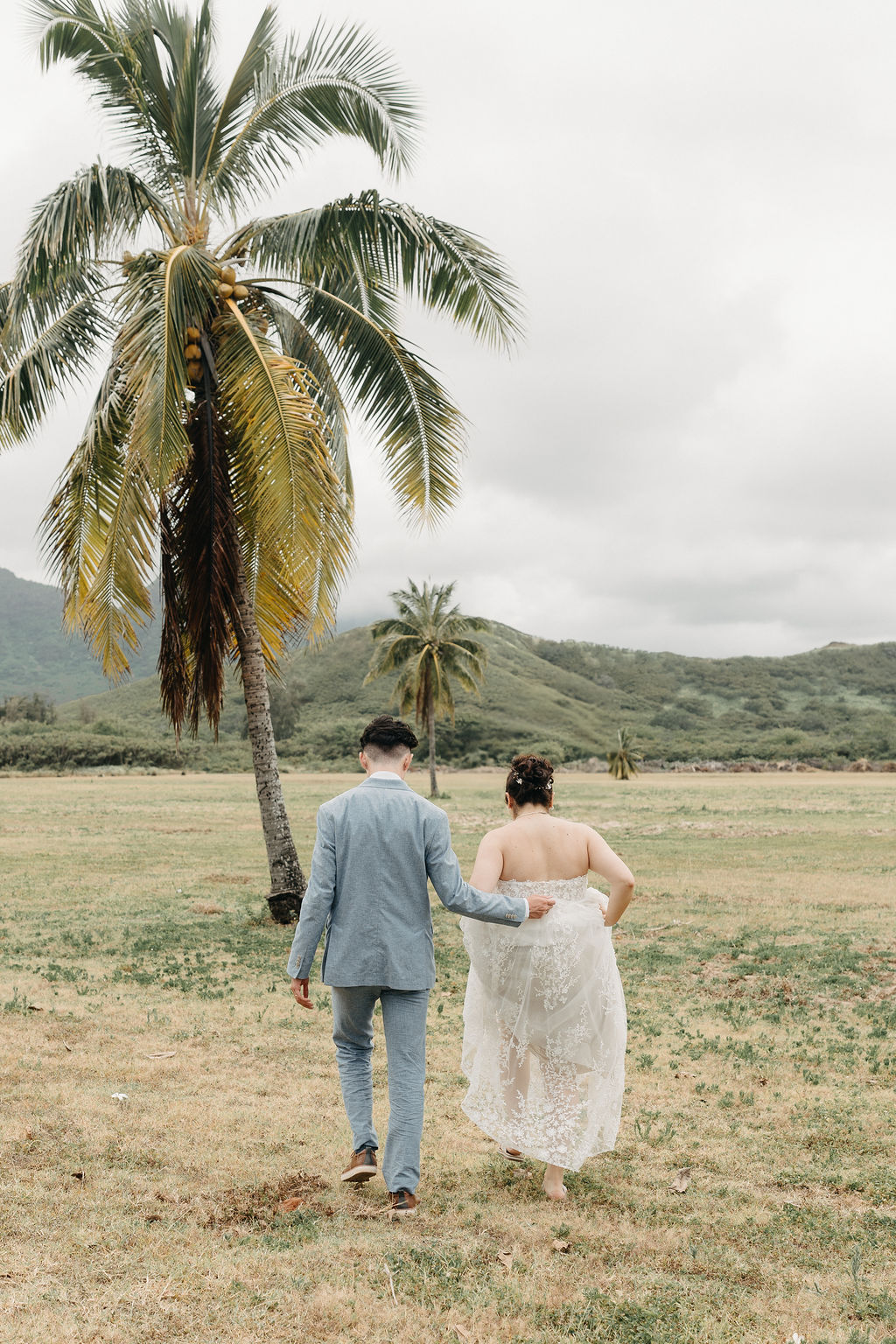 A couple walks hand in hand across a grassy field with palm trees, both wearing formal attire.