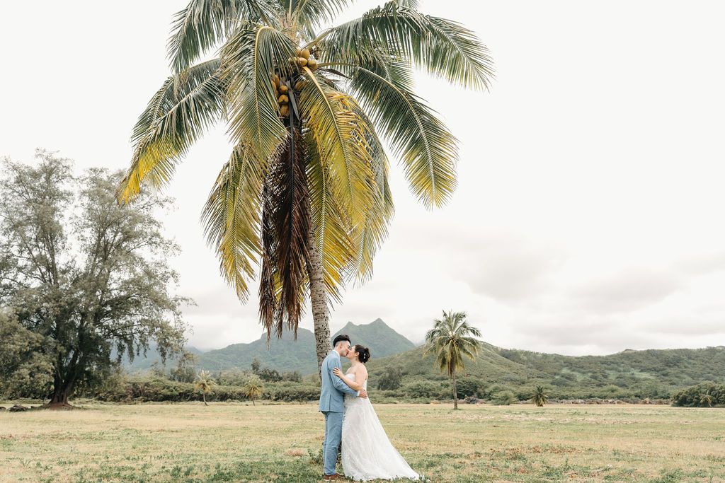 A couple walks hand in hand across a grassy field with palm trees, both wearing formal attire.