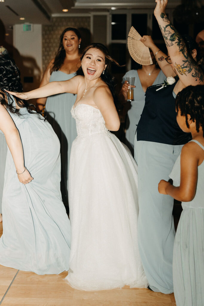 People dancing energetically at a wedding reception with a woman in a white dress leading the dance.