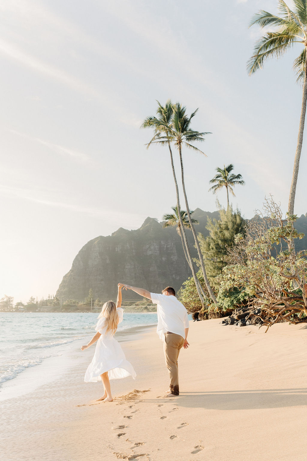 A couple dances on a sunny beach with palm trees and mountains in the background, leaving footprints in the sand.