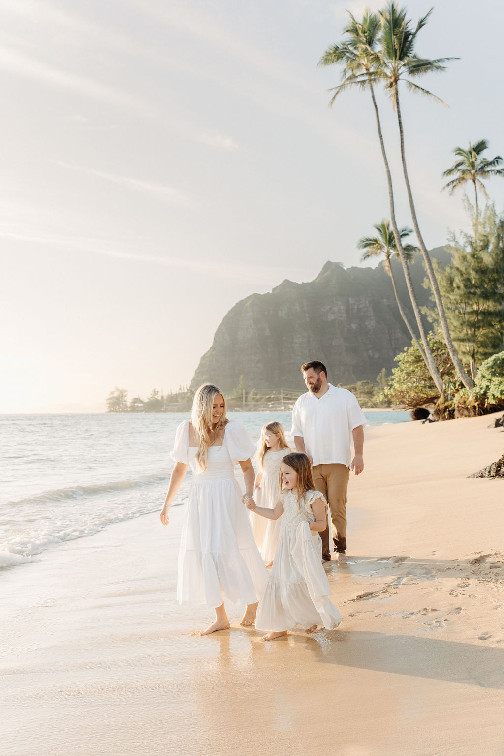 A family in white clothing walks along a sandy beach with palm trees and a mountainous backdrop.