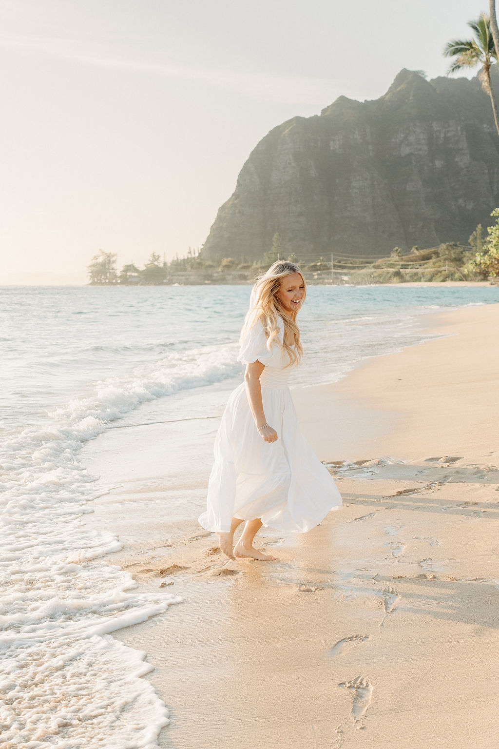 A family in white clothing walks along a sandy beach with palm trees and a mountainous backdrop.