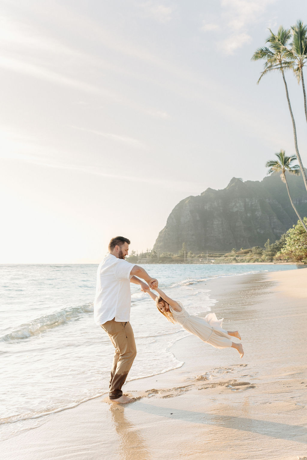 A family in white clothing walks along a sandy beach with palm trees and a mountainous backdrop.