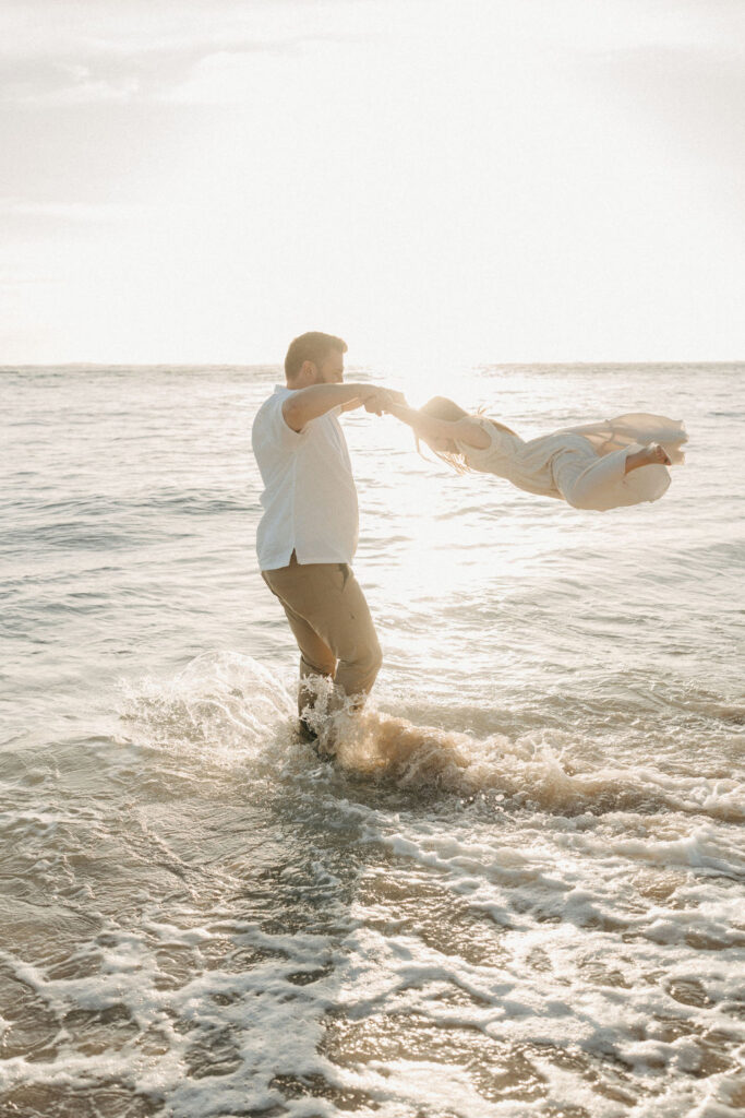 A person stands in shallow ocean water, playfully swinging a child by the arms against a bright sunset.