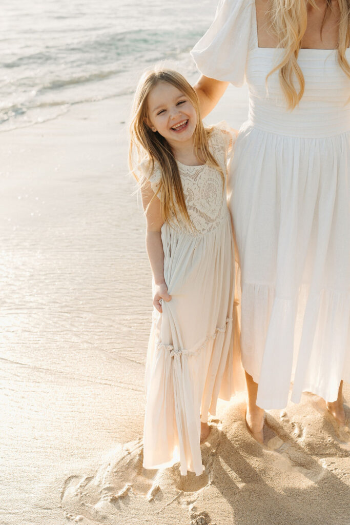 A child in a white dress stands on a sandy beach, smiling, with an adult partially visible beside her in a similar white dress. Waves roll gently onto the shore.