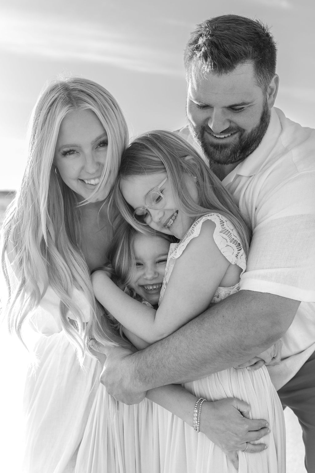 A family of four happily embraces, standing close together. They are outdoors, with the woman, man, and two children all smiling. The image is in black and white.