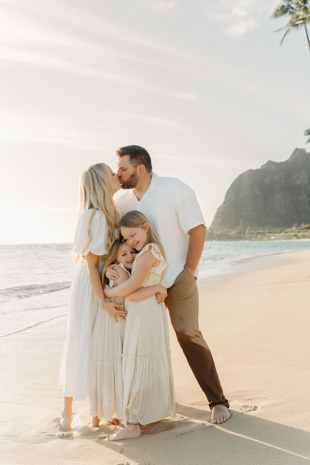 A family of four happily embraces, standing close together. They are outdoors, with the woman, man, and two children all smiling.