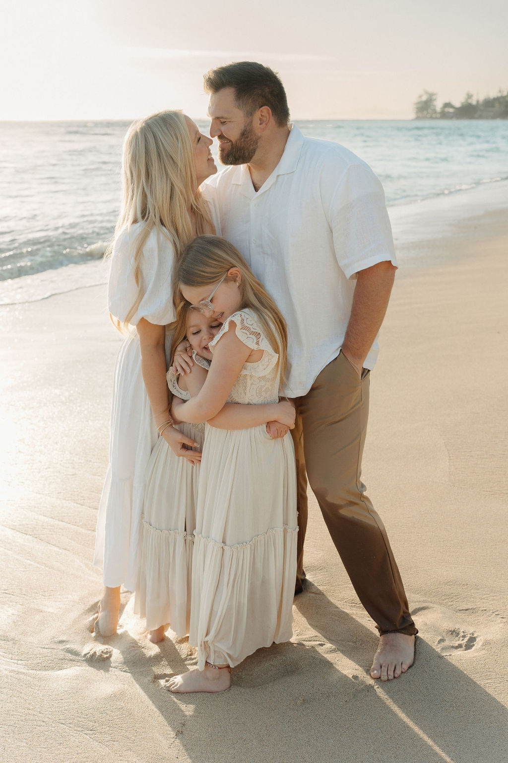 A family of four happily embraces, standing close together. They are outdoors, with the woman, man, and two children all smiling.