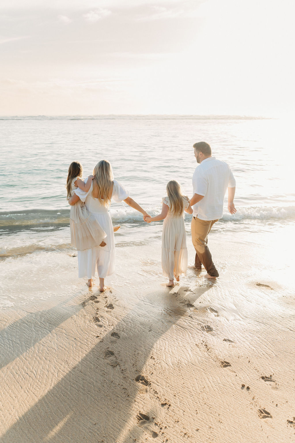 A family of four holding hands walks along the beach, leaving footprints in the sand, with sunlight reflecting on the water.