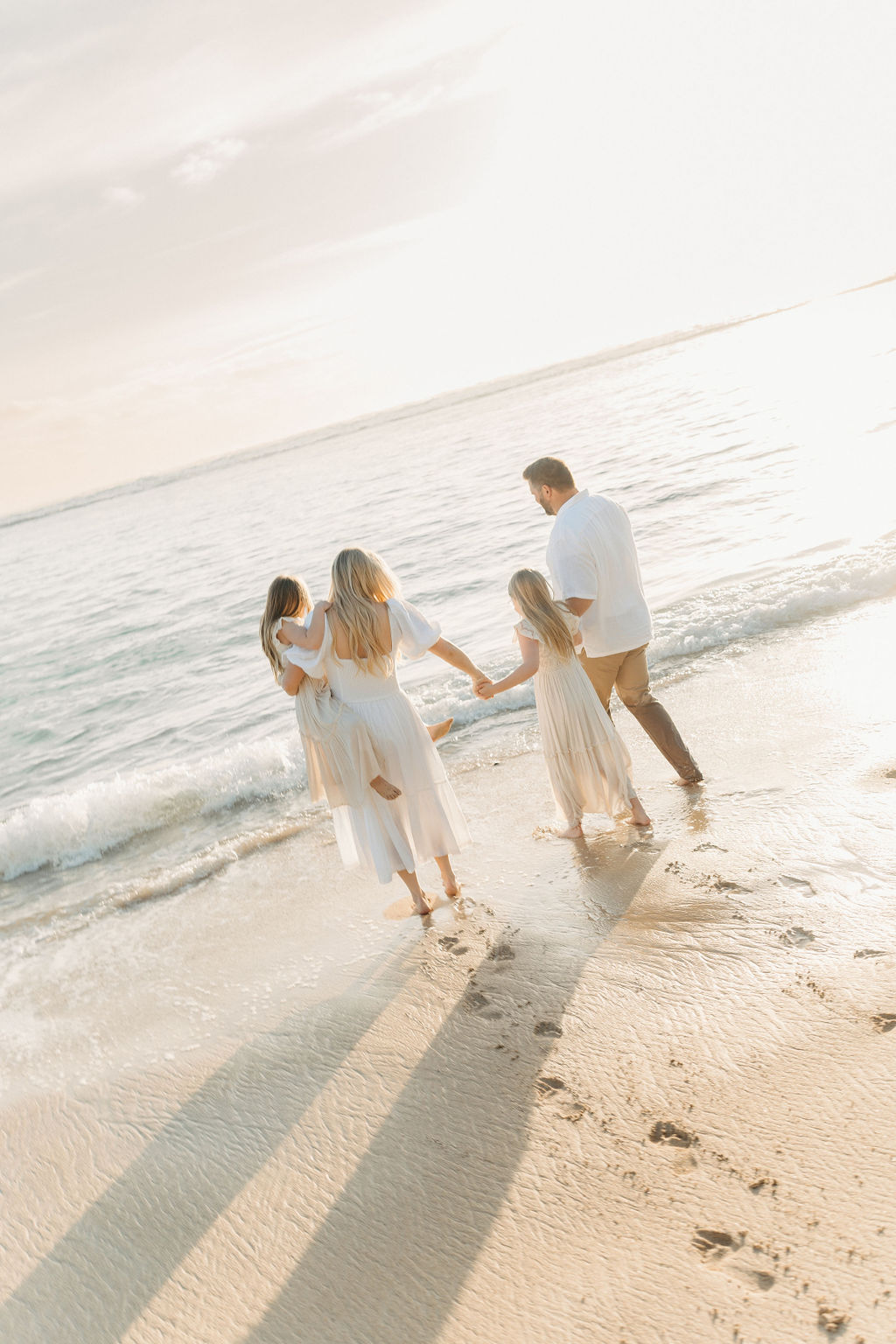 A family of four holding hands walks along the beach, leaving footprints in the sand, with sunlight reflecting on the water.