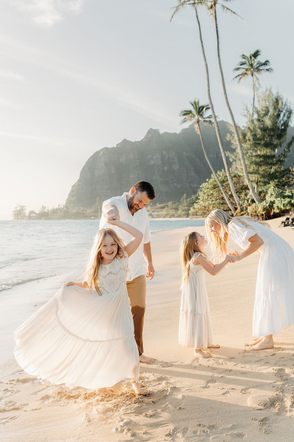 A family of four holding hands walks along the beach, leaving footprints in the sand, with sunlight reflecting on the water.