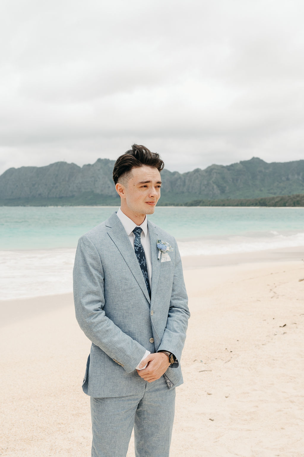 A man in a light gray suit stands on a sandy beach with mountains in the background for a beach wedding in Oahu