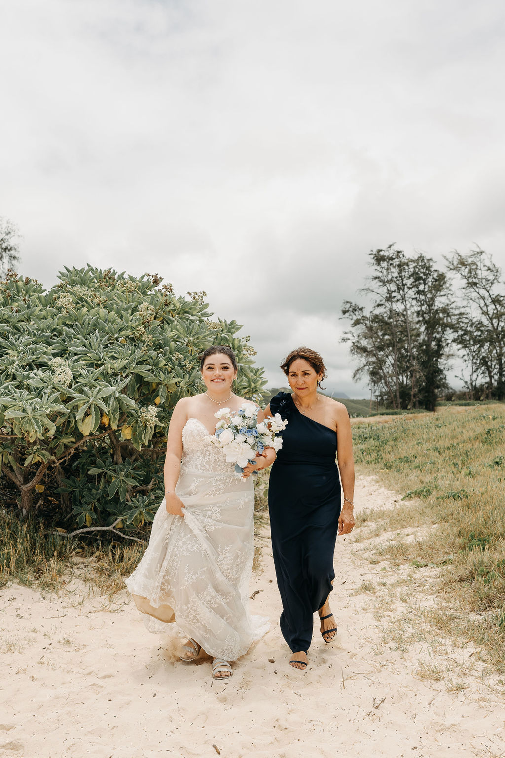 Bride in a white dress holding flowers walks alongside a woman in a dark dress on a sandy path with greenery and cloudy sky in the background.