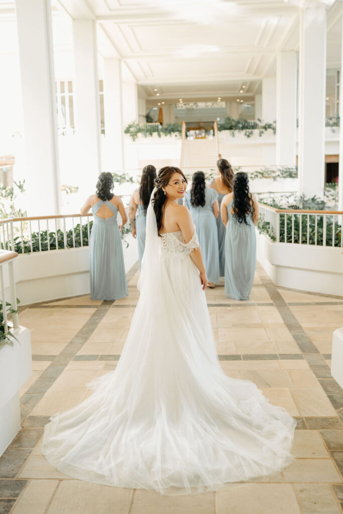 A bride in a white gown with a long train stands indoors, looking back. Six bridesmaids in light blue dresses stand facing away in the background.