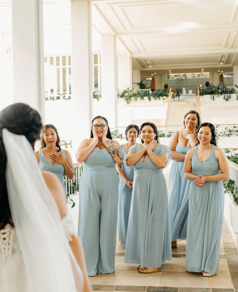 A bride in a white gown with a long train stands indoors, looking back. Six bridesmaids in light blue dresses stand facing away in the background.