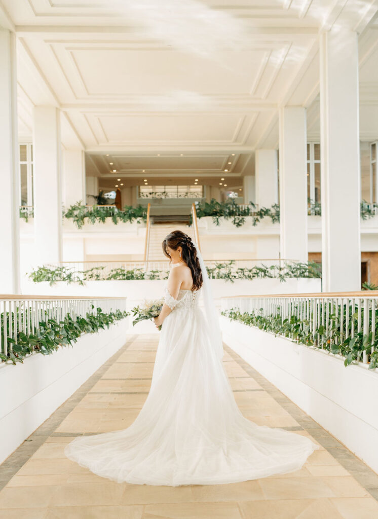 A bride in a white gown holding a bouquet of white flowers stands in a bright, outdoor setting with greenery for her wedding at the four seasons in oahu