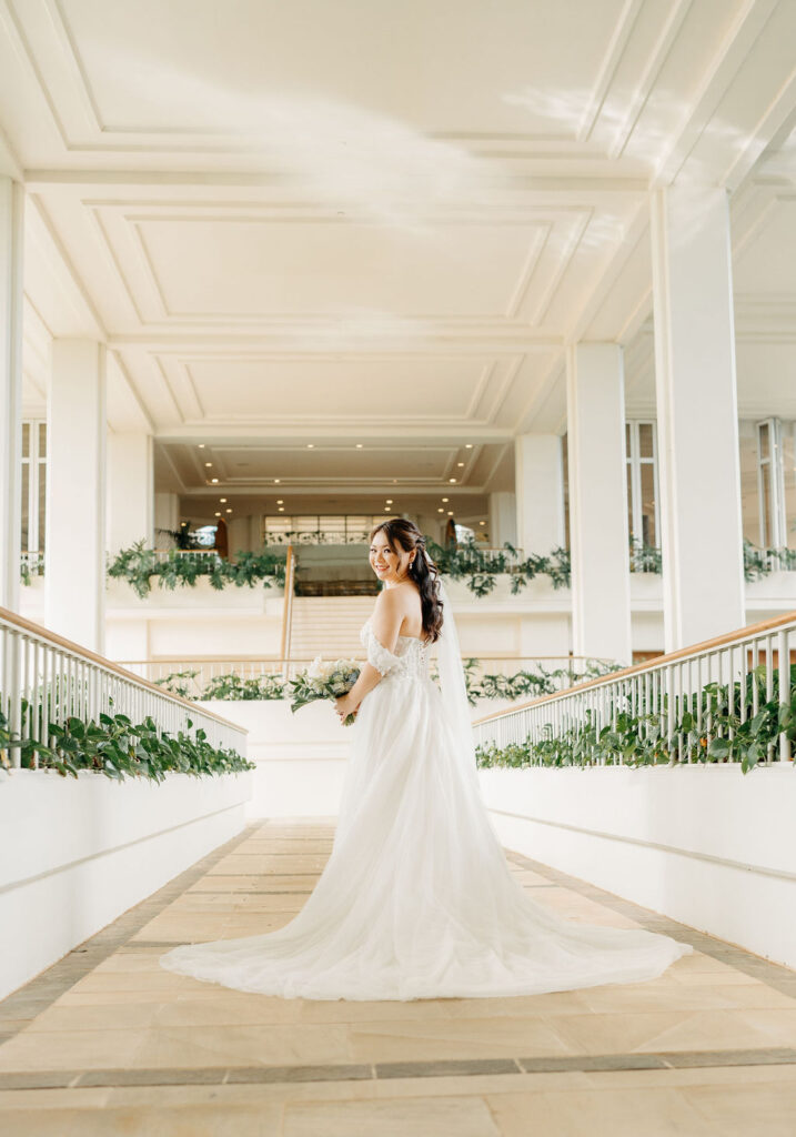 A bride in a white gown holding a bouquet of white flowers stands in a bright, outdoor setting with greenery for her wedding at the four seasons in oahu