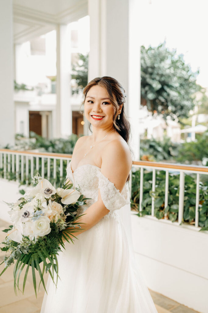 A bride in a white gown holding a bouquet of white flowers stands in a bright, outdoor setting with greenery for her wedding at the four seasons in oahu