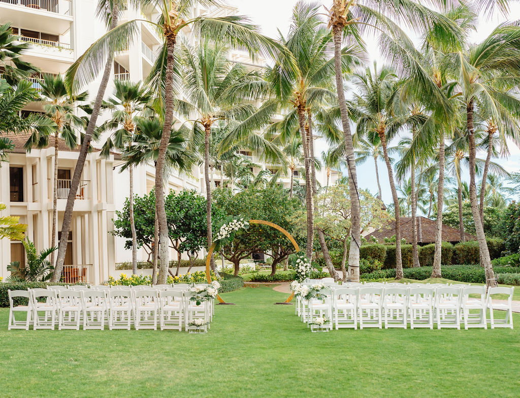 Wedding ceremony setup outdoors with white chairs arranged in rows on a grassy area surrounded by palm trees. An arch decorated with flowers is at the front for a wedding at the four seasons in oahu