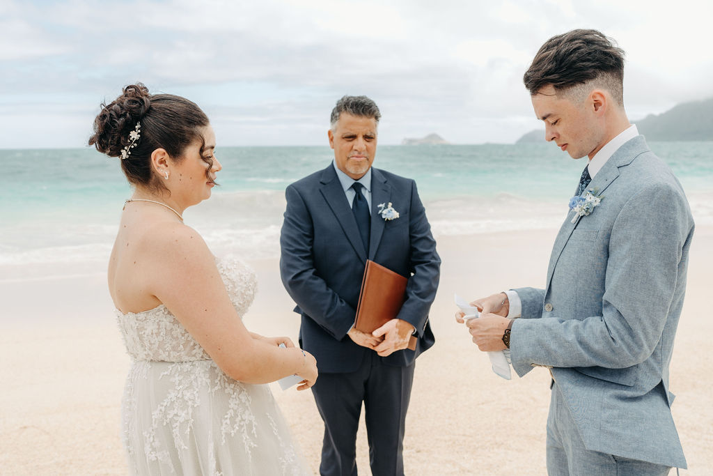 A couple stands on a beach, with the officiant in the middle. The groom reads from a paper. The bride wears a white dress, and the groom is in a light blue suit. The ocean is in the background for a beach wedding in Oahu