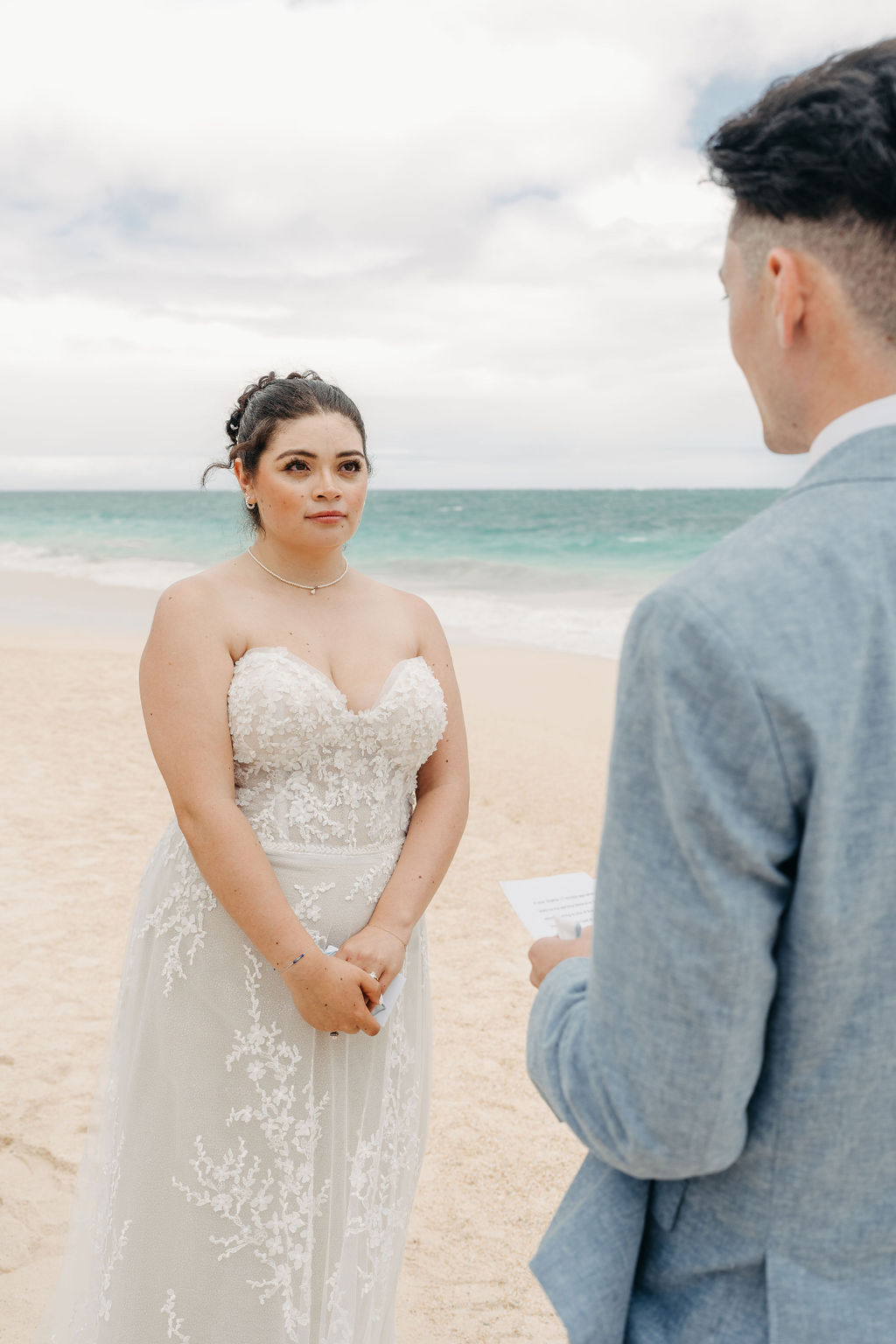 A couple stands on a beach, with the officiant in the middle. The groom reads from a paper. The bride wears a white dress, and the groom is in a light blue suit. The ocean is in the background for a beach wedding in Oahu