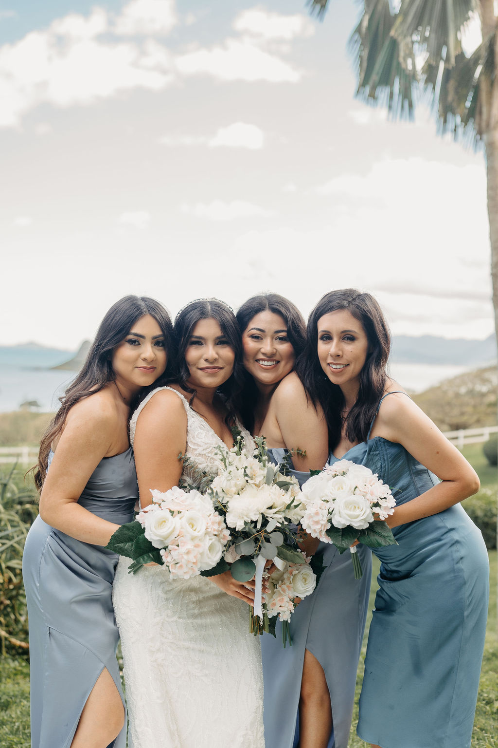 Four women in formal attire pose together outdoors, each holding bouquets of flowers. A palm tree and scenic landscape are visible in the background.