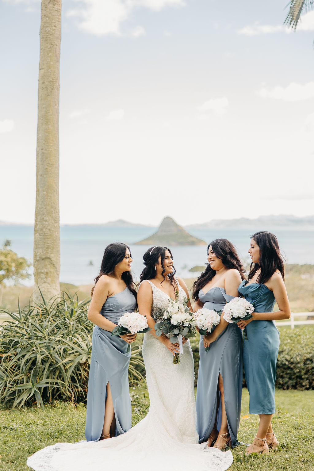 Four women in formal attire pose together outdoors, each holding bouquets of flowers. A palm tree and scenic landscape are visible in the background.