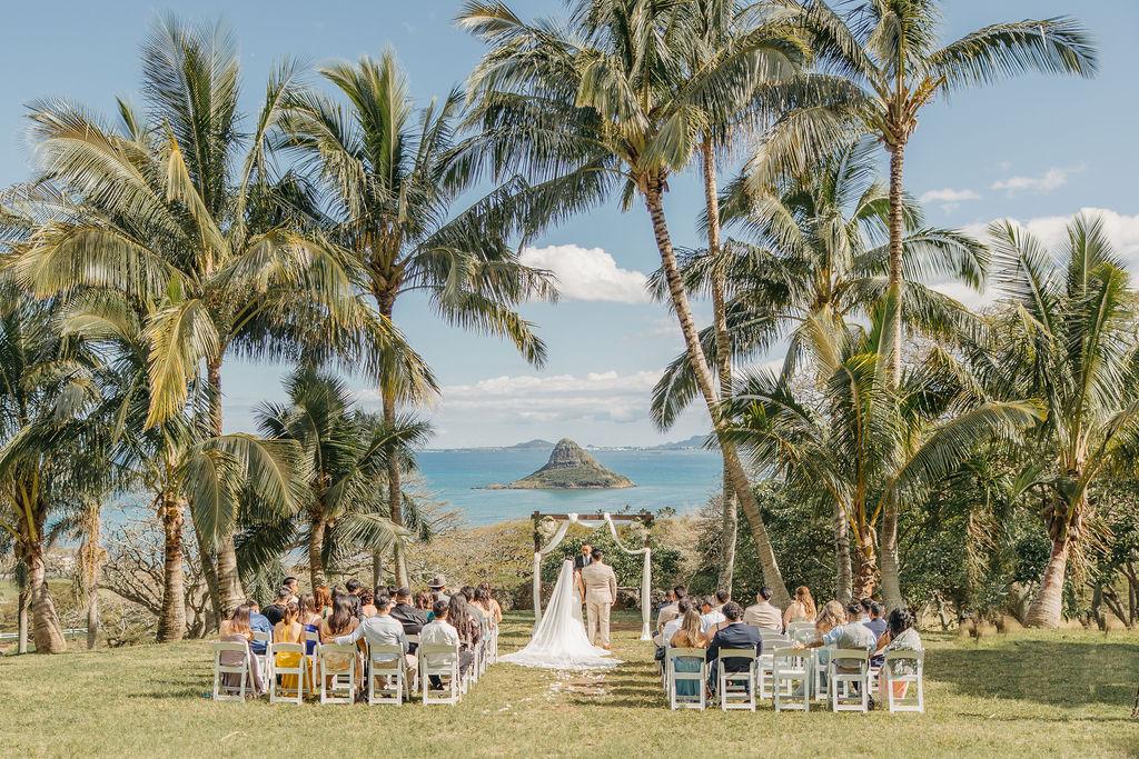 A couple stands under a flower-decorated arch during an outdoor wedding ceremony, with a scenic view of palm trees and a mountain in the background for a destination wedding in oahu hawaii