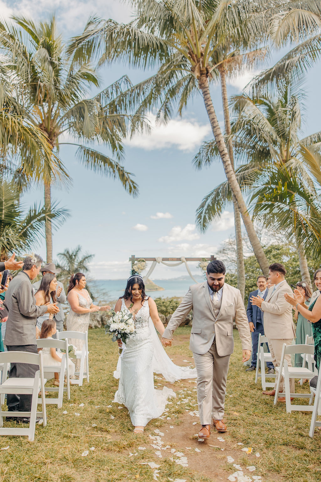 A couple stands under a flower-decorated arch during an outdoor wedding ceremony, with a scenic view of palm trees and a mountain in the background for a destination wedding in oahu hawaii