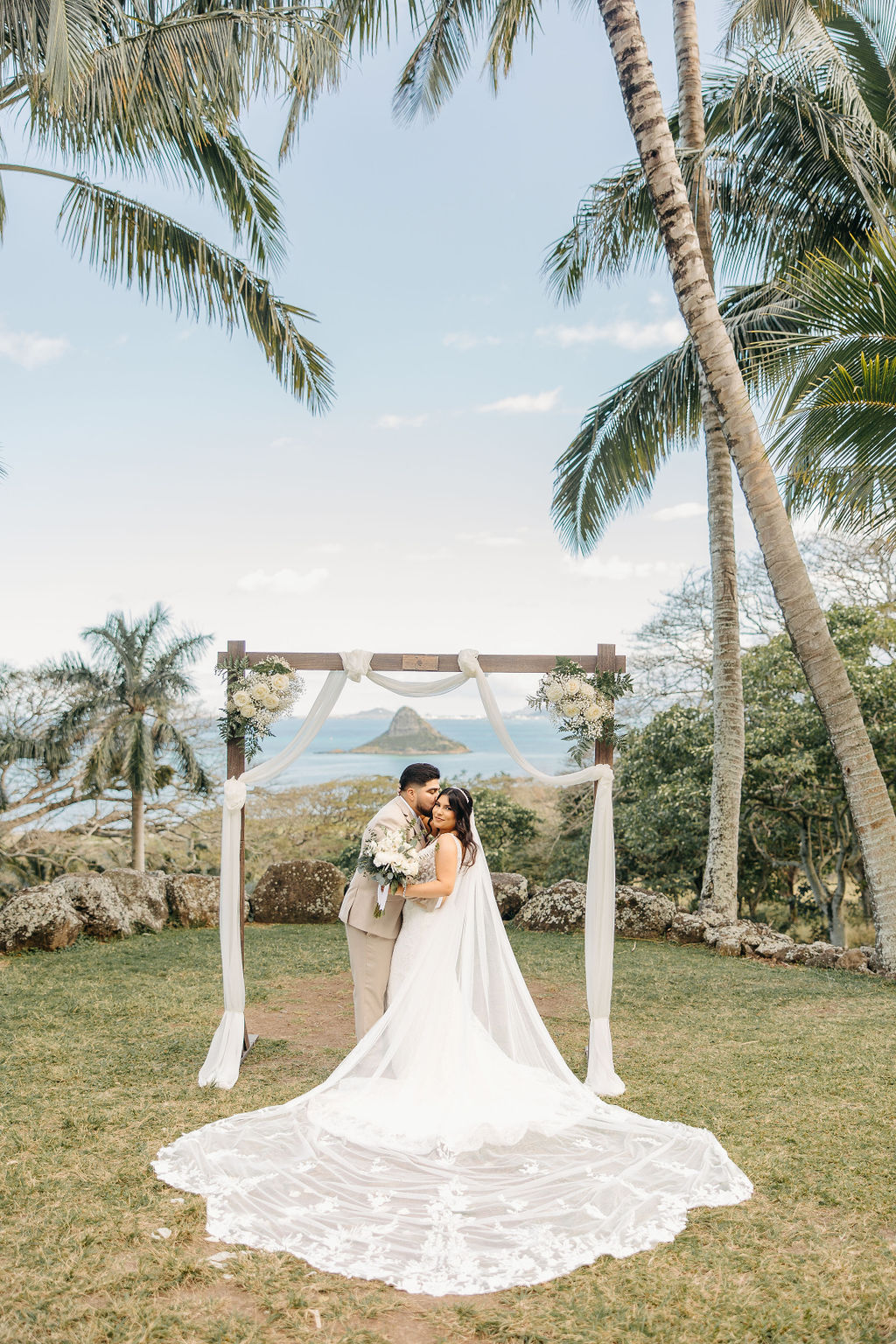 A couple stands under a flower-decorated arch during an outdoor wedding ceremony, with a scenic view of palm trees and a mountain in the background for a destination wedding in oahu hawaii